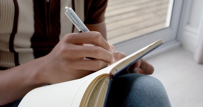 African American man writing in lined notebook by window, pausing, holding clear pen, thinking