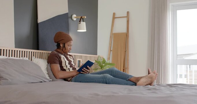 Adult African man holding blue paperback book, reading on king bed, shifting pages glancing window
