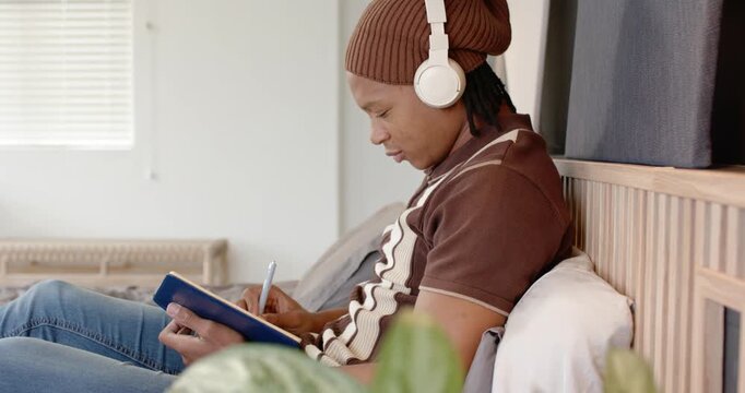 Man sitting on bed with blue notebook on lap starting writing with pen with headphones, journaling