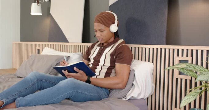 African American man listening and writing in blue notebook on bed, pausing to remove headphones