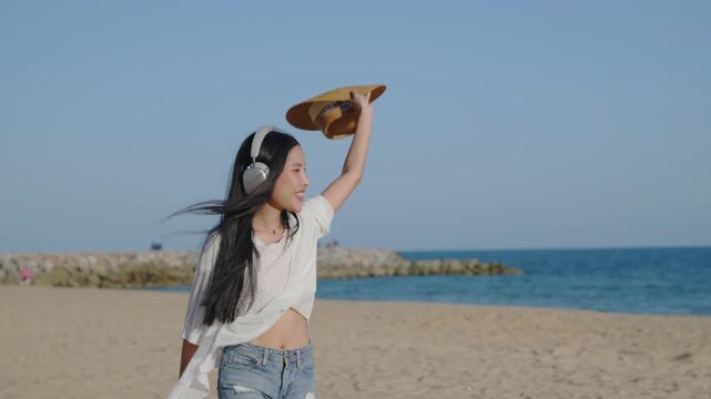 Beautiful happy lady walking on beach by ocean during vacation, wearing wireless headphones and holding straw hat high in air while enjoying sunny summer day under clear blue sky.