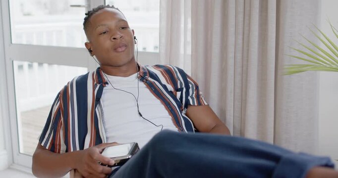 African man in wired earphones reclining near glass door, tuning player and seeking better sound
