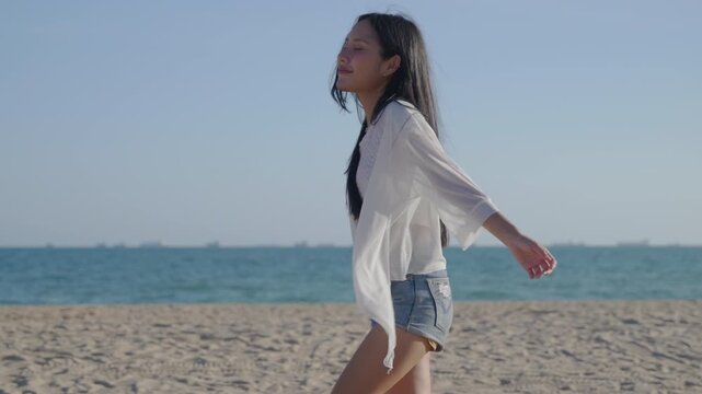 Happy beautiful woman walking on sand of beach enjoying sunset day with freedom and leisure, feeling joyful with arms open wide near calm sea waves during summer vacation.