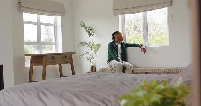 African man in green jacket gazing out window, rubbing brow, thinking on bedroom bench, copy space
