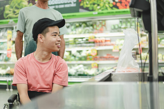 Happy young Asian man in a wheelchair smiling at the weighing scale counter in a supermarket with an assistant, concept of inclusive lifestyle and accessibility.