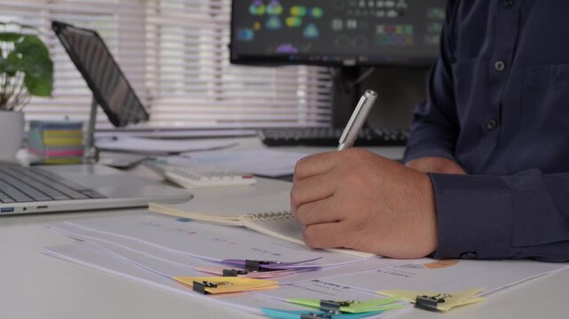 Business professional writing notes and summarizing important information while reviewing charts, reports and documents for data analysis, research, strategic planning and informed decision making