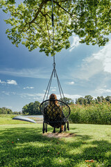 Fototapeta premium Woman seen from behind sits in a hanging chair beneath a leafy tree, overlooking a calm lake with reeds, green lawn and blue summer sky