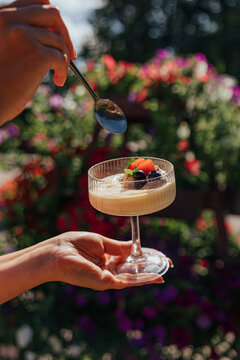 Hands hold a stemmed glass of creamy dessert topped with strawberries and blueberries while a spoon hovers above, with colorful garden flowers softly blurred behind