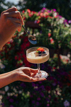 Hands hold a stemmed glass of creamy dessert topped with strawberries and blueberries while a spoon hovers above, with colorful garden flowers softly blurred behind