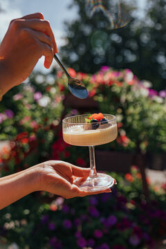 Hands hold a stemmed glass of creamy dessert topped with strawberries and blueberries while a spoon hovers above, with colorful garden flowers softly blurred behind