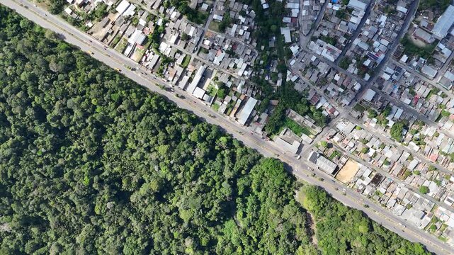 Top-down aerial video of a diagonal street separating dense urban housing from the thick green canopy of the Amazon jungle.