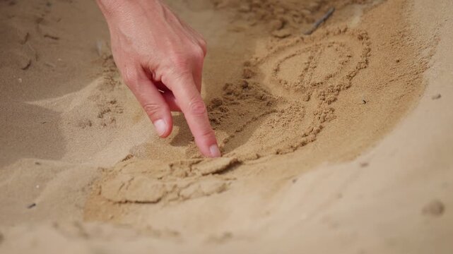 Close-up of a person drawing shapes directly into beach sand with a finger, creating temporary patterns during a relaxing moment outdoors.