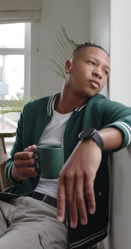 Vertical video: African man in jacket sitting by bright window, glancing at green mug and sipping