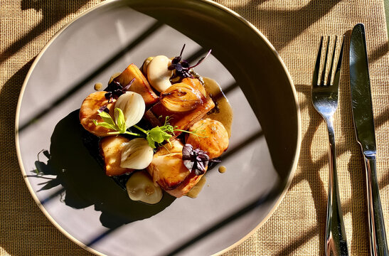 Pork belly served on a modern gray plate with roasted pearl onions, a rich sauce, and fresh herbs, illuminated by harsh sunlight creating strong shadows