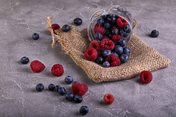 Fresh blueberries and raspberries scattered from a transparent bucket on a gray concrete background © Marina Dubin