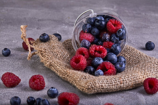 Fresh blueberries and raspberries scattered from a transparent bucket on a gray concrete background