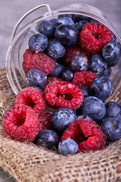Fresh blueberries and raspberries scattered from a transparent bucket on a gray concrete background
