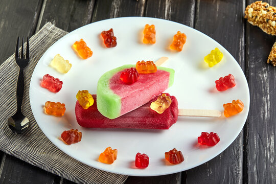 Dessert - ice cream shaped and flavored with watermelon and flavored cherry and candy on a white plate on a black wooden background.