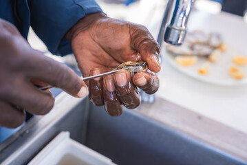 A man opens a fresh raw oyster using a metal shucking knife above a stainless steel sink, with lemon slices visible nearby. © Aerial Film Studio