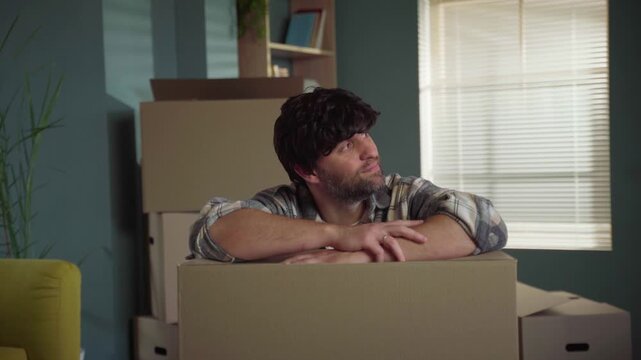 Delighted homeowner leaning on a cardboard box, smiling and looking around his new apartment, enjoying the moment of relocation and envisioning his future life after a successful move