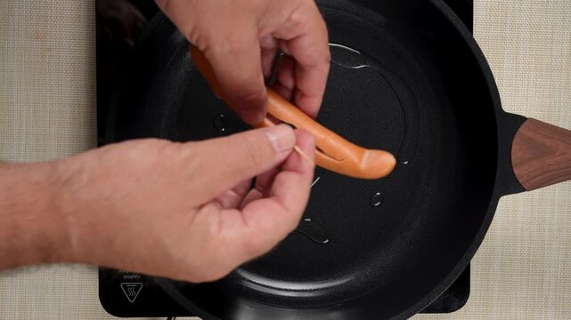 A split sausage is being held open by hand while a wooden toothpick crosses its center. The sausage is positioned inside a black frying pan.