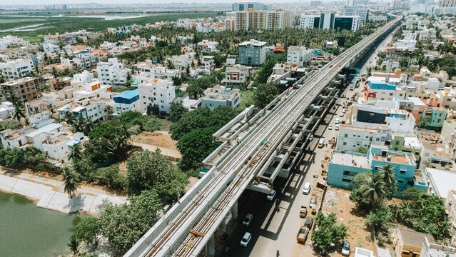 Aerial view of metro rail construction over a busy road in an urban neighborhood with residential buildings, lush trees, and a nearby water body in Chennai, Tamil Nadu, India.