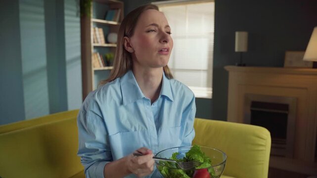 Sad young woman forcing herself to eat a tasteless salad for lunch, showing disgust and displeasure while on a strict diet, sitting on a sofa in her modern living room at home