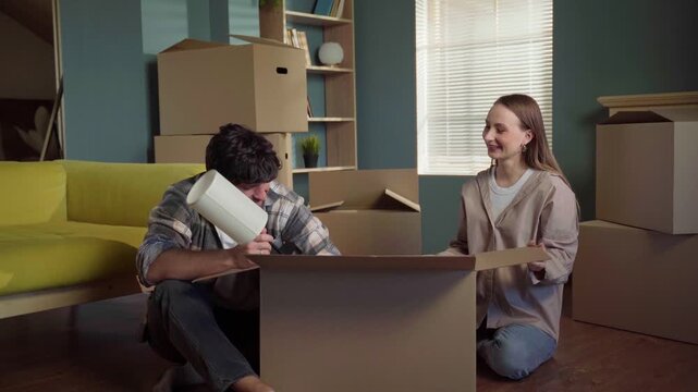 Happy young caucasian couple sitting on the floor in their new apartment, unpacking cardboard boxes and having fun together while assembling a table lamp during their relocation process