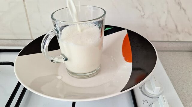 Fresh milk pours into a clear glass mug on a plate beside a kitchen gas stove. Close up indoor footage captures the smooth flow, warm home atmosphere, and everyday cooking scene