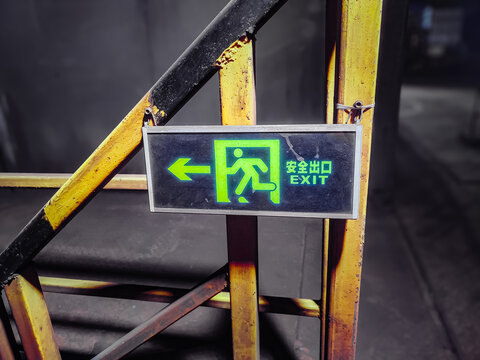 Green emergency exit sign with directional arrow installed on a yellow metal structure in an industrial workplace, representing workplace safety, evacuation procedures, emergency prepa