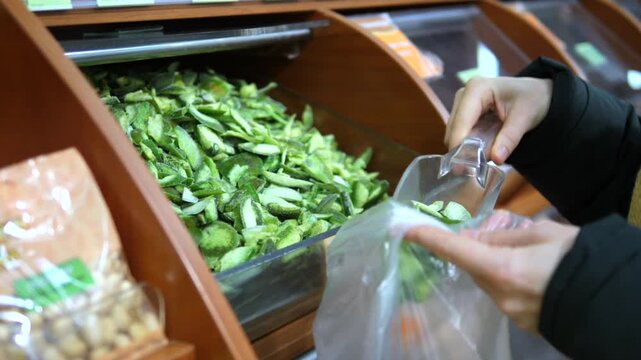 Woman's hand using a plastic scoop to put dried pomelo slices from a self service bulk bin into a bag at a health food store, promoting a zero waste and sustainable shopping experience
