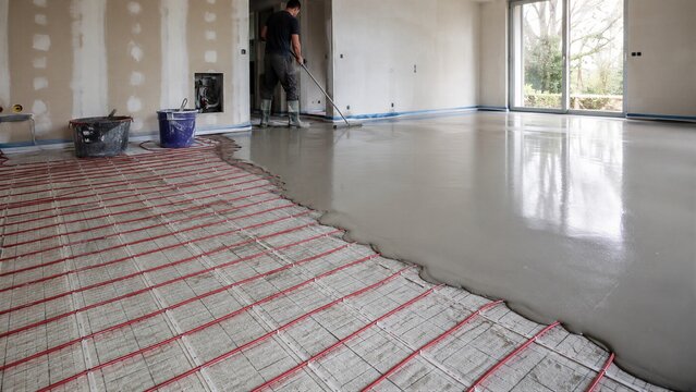 Man worker pouring self leveling screed over red pipe of hydronic underfloor heating system. Warm floor installation and house construction. Energy efficient home heating technology and renovation