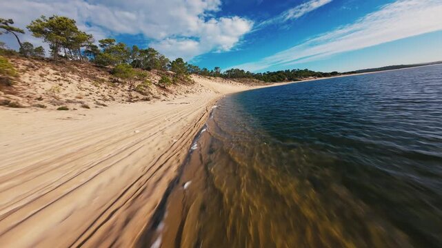 Aerial view of sandy beach with textured dunes and calm ocean waves. Clear blue sky with scattered clouds above. Sand ripples follow waterline under bright sunlight. Scene feels peaceful and natural