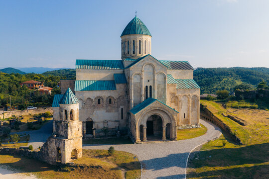 Aerial view of Bagrati Cathedral with its distinctive green dome and stone facade, surrounded by lush green hills and a gravel path in Kutaisi, Imereti, Georgia.