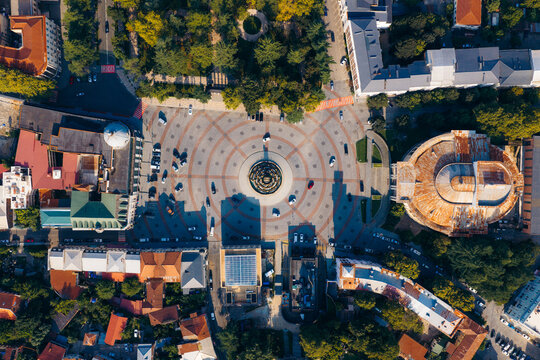 Aerial view of the Colchis Fountain in the center of a circular plaza surrounded by historic buildings, lush trees, and urban streets in Kutaisi, Imereti, Georgia.
