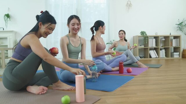 Group asian women in yoga attire and relaxed poses sat together hallway chatting informally. Each sat row on yoga mats corner hall sharing funny stories and snacking on apples friendly manner.