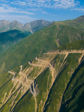 Aerial view of Abano Pass, a winding mountain road carved into steep green slopes with rocky terrain under a clear blue sky in Akhmeta Municipality, Kakheti, Georgia.