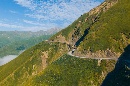 Aerial view of the winding Abano Pass mountain road carved into steep green slopes with rocky terrain and clouds in the valley in Akhmeta Municipality, Kakheti, Georgia.