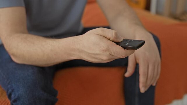 Young hispanic man holding a tv remote control with thumb on button while sitting on orange bed cover and leaning forward in building; relaxation leisure.