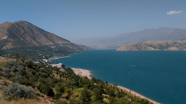 Elevated view of Charvak Reservoir surrounded by dry mountains and green shoreline vegetation under a clear blue sky in Uzbekistan.