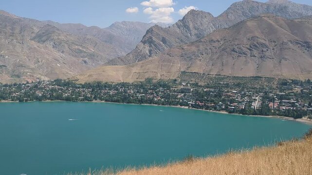 Wide view of Charvak Reservoir with a settlement along the shoreline and rugged mountains in the background, while dry grass sways in the foreground.