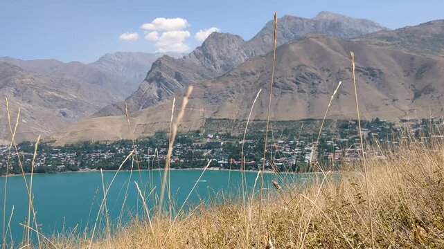 Low hillside view of Charvak Reservoir with dry grass swaying in the foreground, a settlement along the shoreline and rugged mountains in the background.