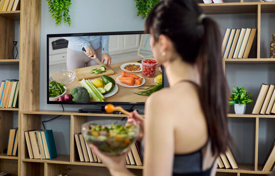 Woman eating salad while watching television. She watches a cooking show and enjoys fresh vegetables at home, supporting healthy diet and fitness lifestyle. Wellness and healthy living concept.