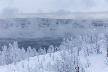 Scenic winter view of a steaming, ice-free bay surrounded by snow-covered hills and frost-covered birch trees in arctic cold. © AndreyZayats