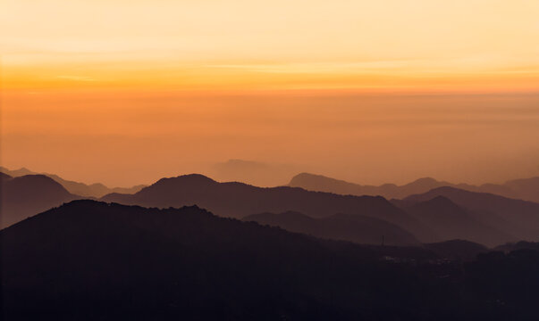 Aerial view of the layered mountain silhouettes of Telaga Dringo at sunset with hazy atmosphere and golden sky in Indonesia.