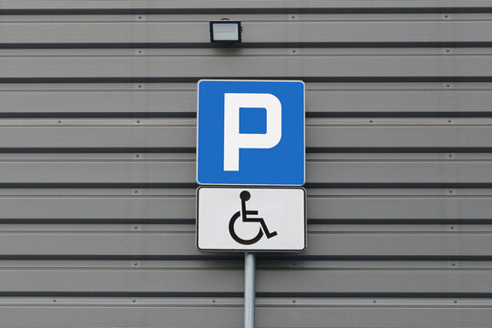 Accessible parking sign marks reserved vehicle space. Wheelchair symbol identifies parking for disabled drivers. Blue sign stands against metal wall. Public accessibility parking background.