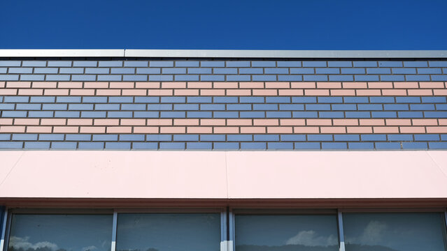 striking building facade with a vivid blue sky backdrop, striped gray-terracotta brick tiles, and a soft pink base wall framing dark glass windows below