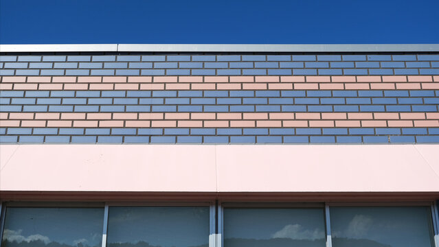 striking building facade with a vivid blue sky backdrop, striped gray-terracotta brick tiles, and a soft pink base wall framing dark glass windows below