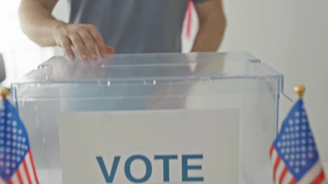 Man inserting paper ballot into a transparent ballot box with hand visible, american flags nearby inside a public building; patriotic civic duty.