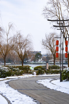 Snowy park walkway beside a river with bare trees and red lantern decorations